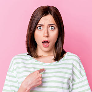 Young brunette woman with shocked open mouth expression pointing to herself in front of pink background