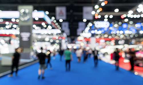 Blurry photo of a tradeshow floor with people walking, spots of lights, booths, and blue carpet